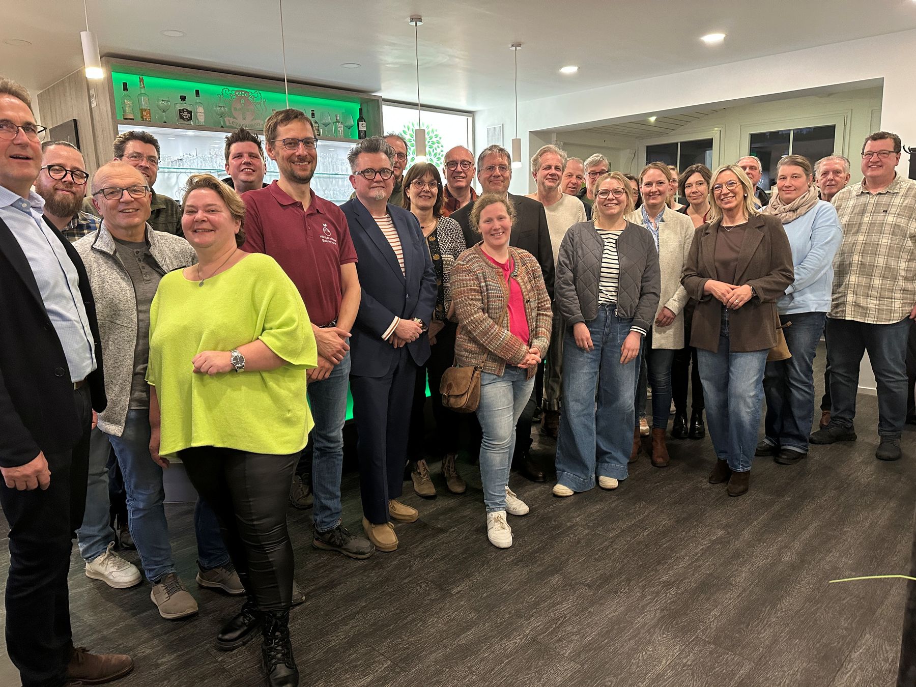 Eine Gruppe von etwa 25 Erwachsenen, die in einem Innenraum in ungezwungener Atmosphäre stehen und in die Kamera lächeln. Im Hintergrund ist ein Barbereich mit Getränken und Regalen zu sehen, und alle sind leger bis elegant gekleidet.